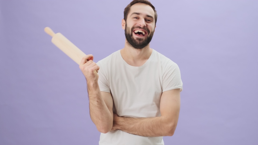 A happy joyful young man wearing a white t-shirt is holding a rolling pin standing isolated over gray background