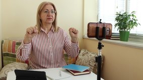 Mature woman shows finger gymnastics through a video call on a smartphone. Learn over the Internet, records assignments. Record a video for blog about health. Online education for adults.