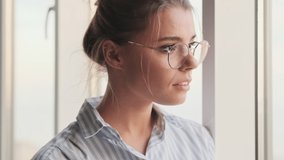 A beautiful calm young woman is putting off her glasses while looking out the window standing at home in the living room