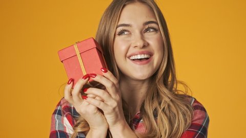 Young cheery girl isolated over yellow wall background holding present box trying to guess what's inside