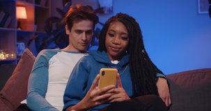 Front view of young happy multiethnic couple looking at smartphone screen while sitting on sofa. Millennial boyfriend and girlfriend browsing internet while spending time together at home.