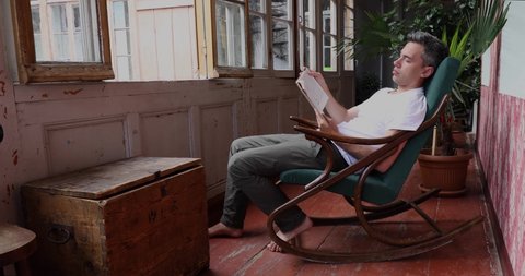 Young man sitting and reading in rocking chair in the old rural house. The guy is relaxing while slowly rocking in her old armchair with book in his hands