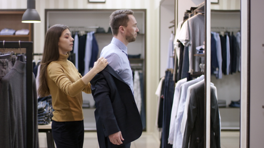 A side view slow motion video of a young female clothing boutique consultant helping a male client put on a jacket to see how it looks on him | Shutterstock HD Video #1052749811