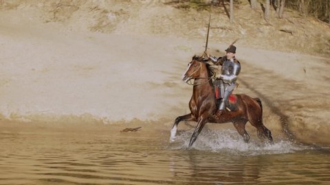 Video footage, a young adult man in knightly armor rides a horse on water in a river along a sandy shore