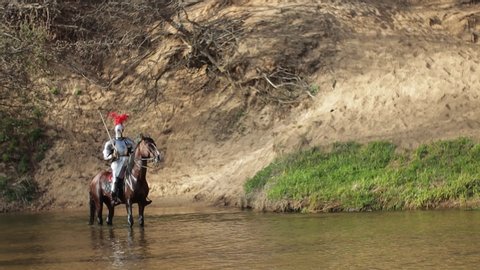 Video footage, a young adult man in knightly armor rides a horse on water in a river along a sandy shore