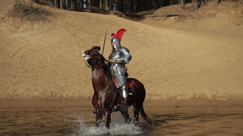 Video footage, a young adult man in knightly armor rides a horse on water in a river along a sandy shore Royalty-Free Stock Footage #1052750333