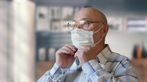 Concept of health and safety of old people during the pandemic of the coronavirus COVID-19. Elderly man with glasses dressing up a handmade homemade protective mask indoors