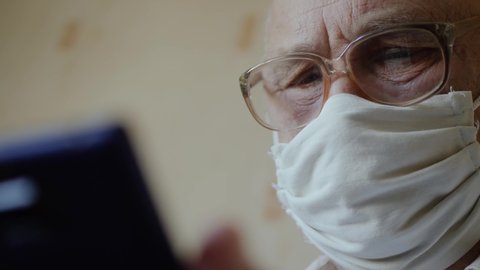 Sad elderly man in isolation at home wearing a handmade virus protection mask uses phone close up. Reading news from a smartphone in a home interior. Old man with glasses stays in quarantine at home