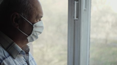Elderly man in a protective mask on his face looks out the window copy space. Quarantine Restriction Concept. Old man indoors during an epidemic