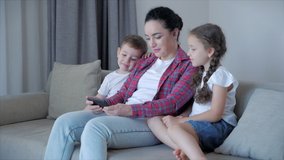 Happy family,mom and cute little children watch the phone,mom plays with children at home,relaxing using a smartphone,hugging,sitting on the couch, laughing, having fun,enjoying moments of family life