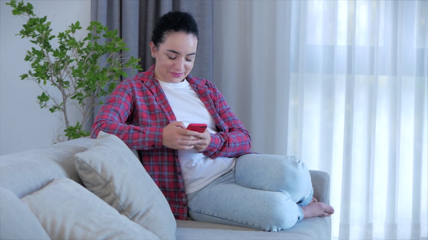 Woman typing on phone sitting on sofa at home, businesswoman sits at home types on smartphone checks mail to study online work at home. Royalty-Free Stock Footage #1052762354