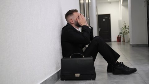 Fired business man sitting frustrated and upset in the hallway near office with his bag. He lost work