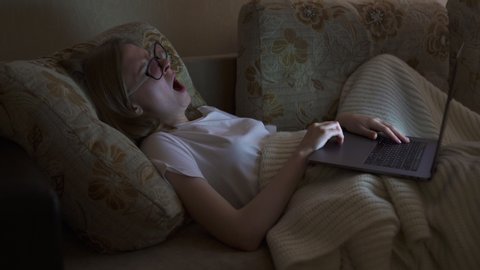 Young Girl lying on a sofa with a laptop and yawns few times, then falls asleep. Trying to work at night. Girl with long hair and wear a white shirt. Legs are covered with a plaid.
