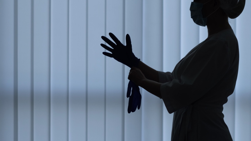 Young women doctor in medical suit and cap putting on rubber gloves in hospital or clinic on the background of a large window. Medium shot silhouette | Shutterstock HD Video #1052779922