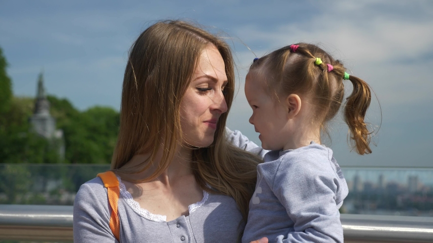 Loving Mother Kisses Child While Holding on Hands on Bridge. City Background Bright Sunny Day