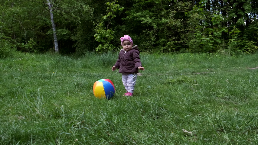 A baby girl plays with a multi-colored inflatable ball on the lawn in the park.