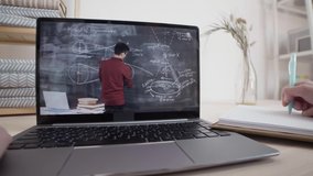 Point of view shot of unrecognizable person sitting in front of laptop, watching young Asian male teacher drawing formula on blackboard and making notes in notebook lying near laptop