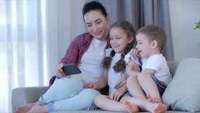 Happy family,mom and cute little children watch the phone,mom plays with children at home,relaxing using a smartphone,hugging,sitting on the couch, laughing, having fun,enjoying moments of family life