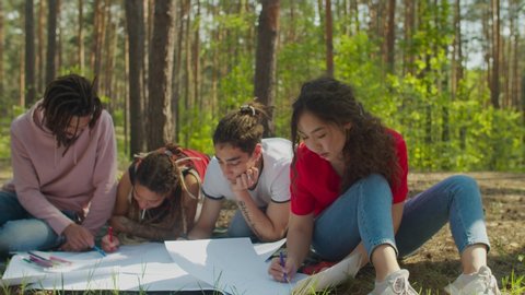 Team of diverse multiracial college students creating ecological project together in forest. Eco-friendly multiethnic volunteers making placards against environmental pollution outdoors.
