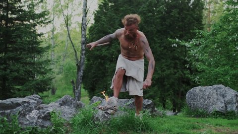 Comic Primitive Savage Making Fire in Forest and suddenly Burning his Fur Shoes. Shocked Naked, wild Caveman Set a Fire. Evolution Process, Prehistoric stone age. Slow Motion.