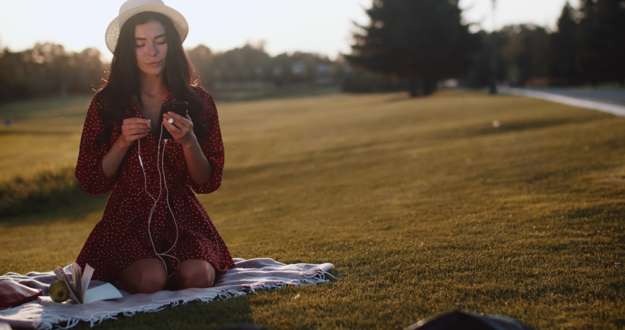 Smiling girl in a red dress with a phone in her hands listens to music at sunset in the park. A young woman is sitting on the grass with a phone and headphones 