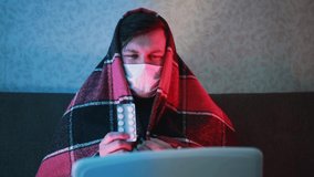 A man wrapped in a blanket and in a mask holds a pack of tablets in his hand and consults via video link on a laptop.