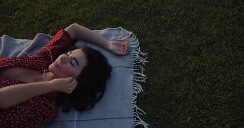 Beautiful happy girl in a red dress listens to music at sunset in the summer park. Young woman with a phone and headphones. Lovely girl enjoying music while lying on the grass Video de stock