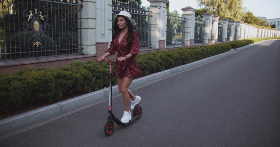 Beautiful girl rides a scooter in the summer park in slow motion. Cute woman in a dress roll and smiles. Young female is engaged in a hobby outdoors 