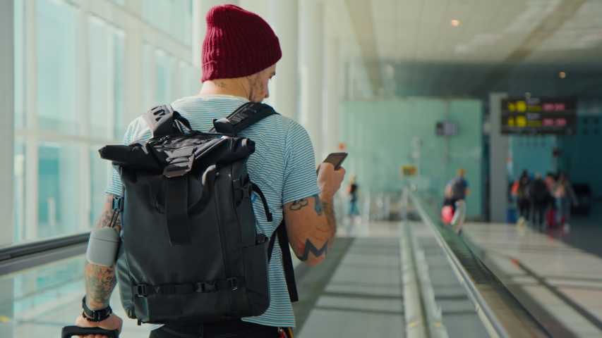 Casual trendy young man with backpack and suitcase travels home through big empty airport. Male traveler look at phone, search for boarding pass or flight cancelation information. Airport closure