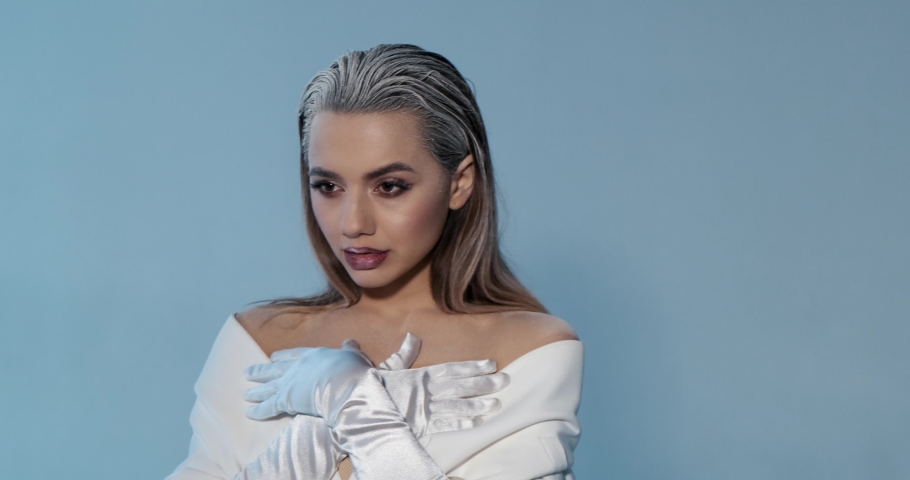 Beautiful young model poses for camera in photo studio, folds hands, looks up, poses, winter makeup, white hair, gloves, open dress, cherry lips, winter queen, magazine cover, girl with catchy makeup