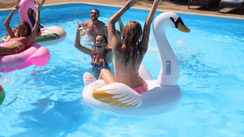 Friends have party in a private villa swimming pool. Happy young people in swimwear splashing water, dancing with floaties and inflatable mattress in luxury resort on sunny day. Slow motion. | Shutterstock HD Video #1052804396