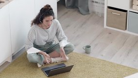 High angle view of Caucasian woman wearing headset sitting on kitchen floor with laptop on her knees, having video chat, drinking coffee then looking at camera and smiling