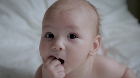 Portrait of a nice newborn baby in diaper , lying on his stomach in a white bed sucking fingers, looking interestingly at camera with his mouth open and legs dangling. Slow motion