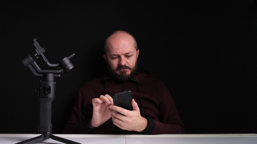Videographer in hoodie sitting behind the desk with new axis gimbal stabilizer, typing on smart phone