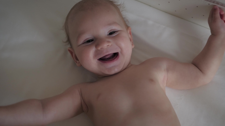 A nice little caucasian newborn baby is funny smiling, lying at the back in the child crib. Portrait of a playful and energetic child close-up, slow motion