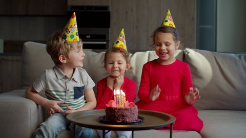 Three kids in birthday hats celebrating birthday with chocolate cake with candles at home. Girl blowing out birthdays candles on cake with siblings sitting on couch in living room. 