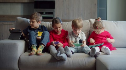 Group of four mixed aged kids using gadgets sitting together on couch at home. Siblings playing and watching online on mobile phones and tablet computers. Two boys and two girls with devices. 