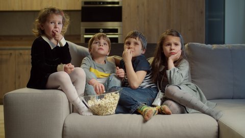 Group of four kids watching TV, eating popcorn sitting on couch at home. Two girls and two boys watching exciting movie at home. Siblings on couch. 