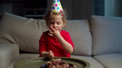 Cute little girl in party hat eating birthday chocolate cake with hands at home. Birthday kid enjoying eating sweets alone at home. 
