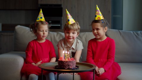 Three kids in birthday hats celebrating birthday with chocolate cake with candles at home. Brother blowing out birthdays candles on cake with sisters sitting on couch in living room. 