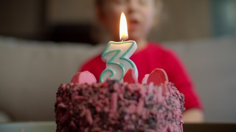 Close up of kid blowing out candle with number 3 on chocolate birthday cake in slow motion. Three years old girl celebrates birthday. 
