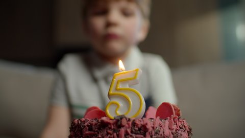 Close up of kid blowing out candle with number 5 on chocolate birthday cake in slow motion. Five years old boy celebrates birthday. 