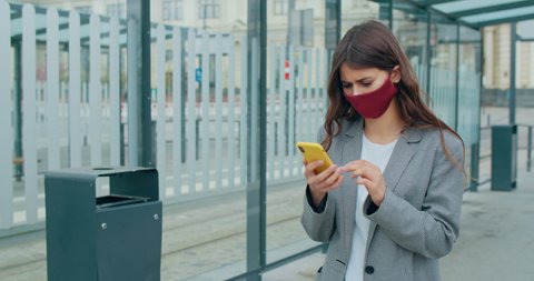 Woman in reusable face mask browsing internet or searching job. Millennial girl using her smartphone and scrolling and touching screen while walking at city street. Concept of COVID