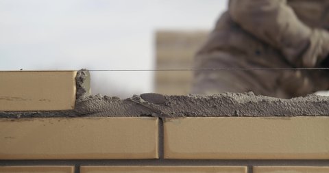 Close-up of construction work, male bricklayer puts another yellow brick into mortar, neatly smooths edges with trowel, taps, works well, professional, building private house from brick, construction