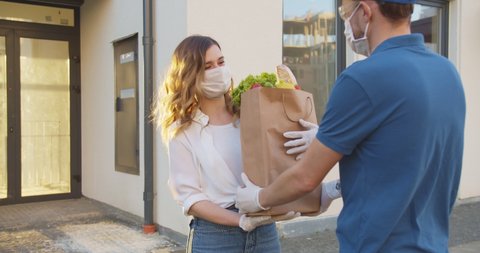 Side view of delivery man walking from supermarket coming to building and meeting young Caucasian woman. Girl in medical mask and gloves handling packet with food from courier man. Pandemic concept.