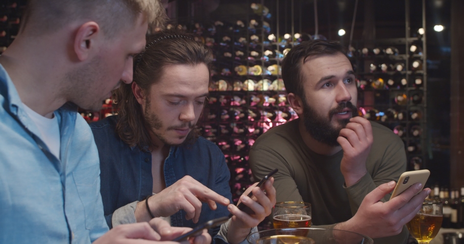 Caucasian male young handsome football fans watching match in pub and sending messages from mobile phones. Guys placing bets on sport game result. Men raising rates of bookmakers by cellphones in bar. | Shutterstock HD Video #1052831060