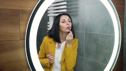 Appealing serious 30-aged stylish woman with black hair dressed in olive colour suit, putting pomade on her lips while looking in the mirror before exit from home