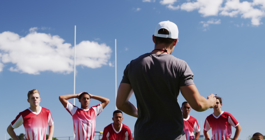 Rear view of a Caucasian male rugby coach standing on a playing field holding a clipboard talking to teenage multi-ethnic male team of rugby players wearing their team strip standing in slow motion Royalty-Free Stock Footage #1052833754