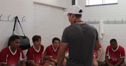 Rear view of a Caucasian male rugby coach standing in the changing room talking to a teenage multi-ethnic male team of rugby players wearing their team strip, sitting and listening