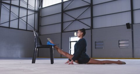 Side view of teenage mixed race female gymnast practicing at the gym, doing splits and stretching with one leg on a chair. Gymnast training hard for competition in slow motion.